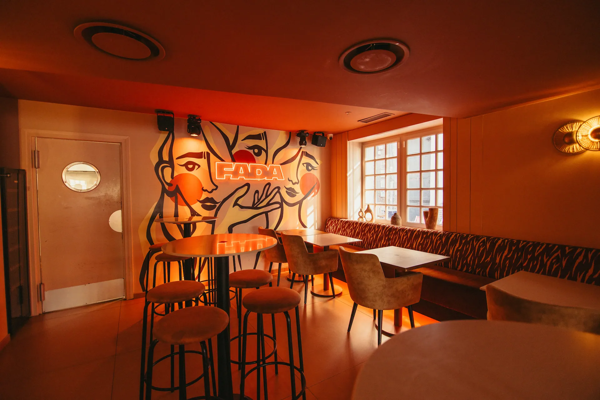 La salle du bar, tables hautes et tabourets devant une fresque de visages signée FADA, les fenêtres à droite