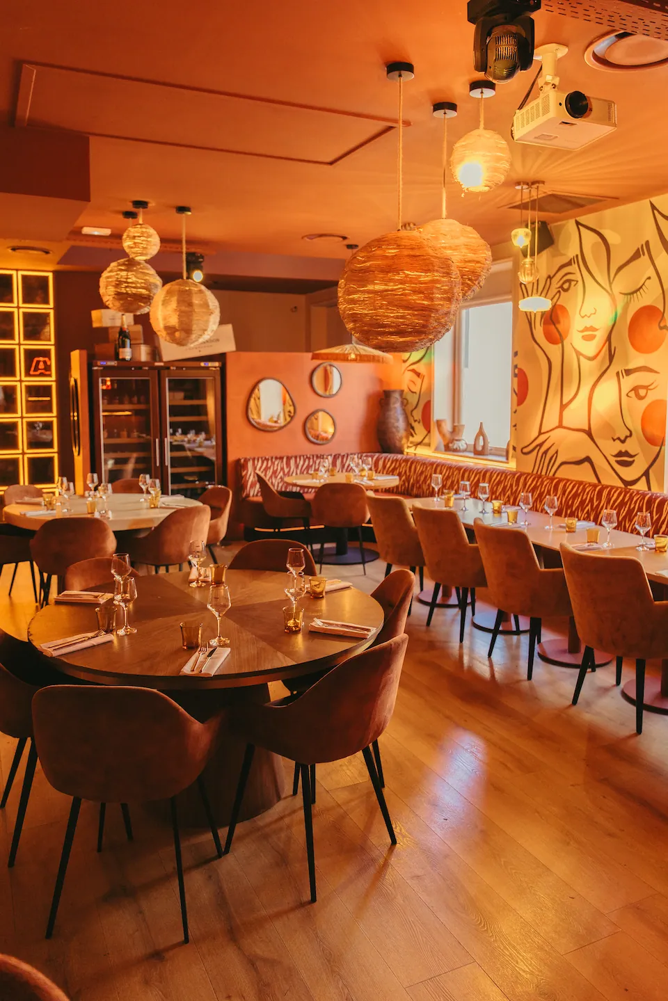 La salle du restaurant baignée de lumière chaude, tables rondes dressées sous des suspensions en osier et une fresque de visages au mur