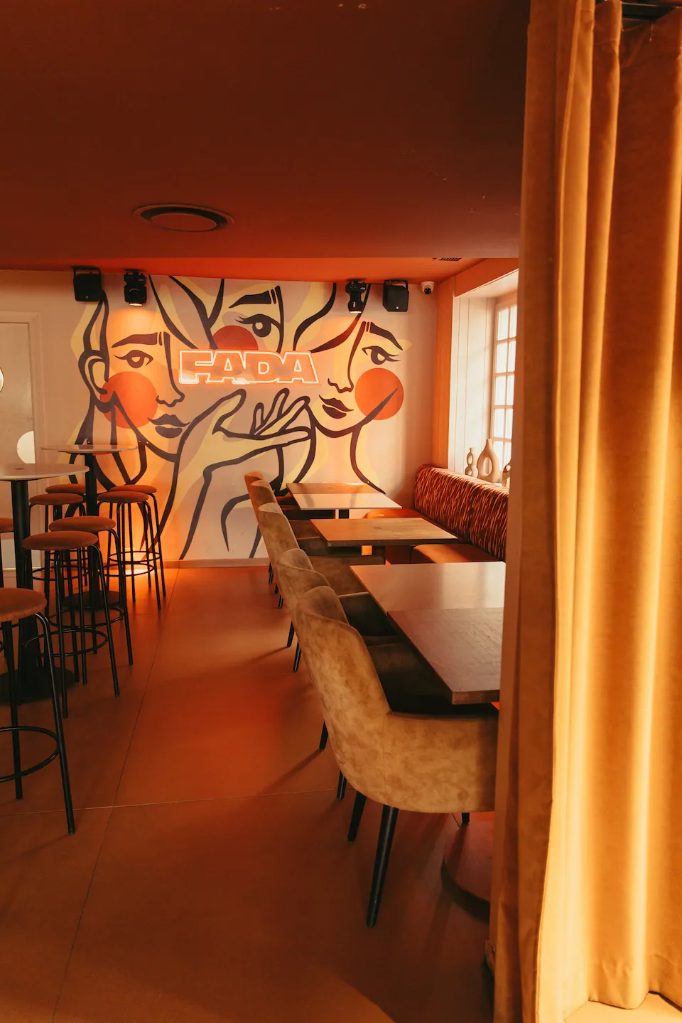 Une salle aux grandes tables et banquettes, devant une fresque de visages signée FADA, un rideau doré au premier plan