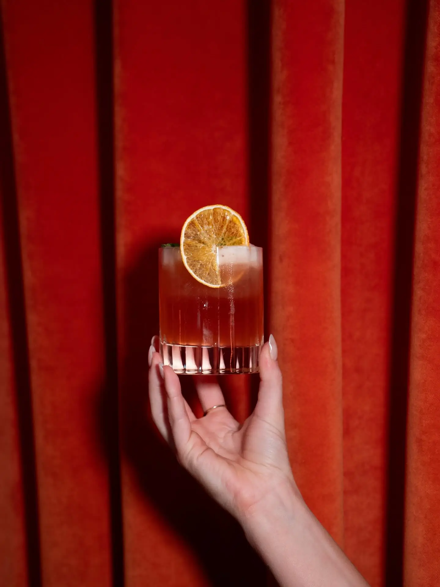 Un cocktail servi dans un verre strié, tenu devant un rideau de velours rouge